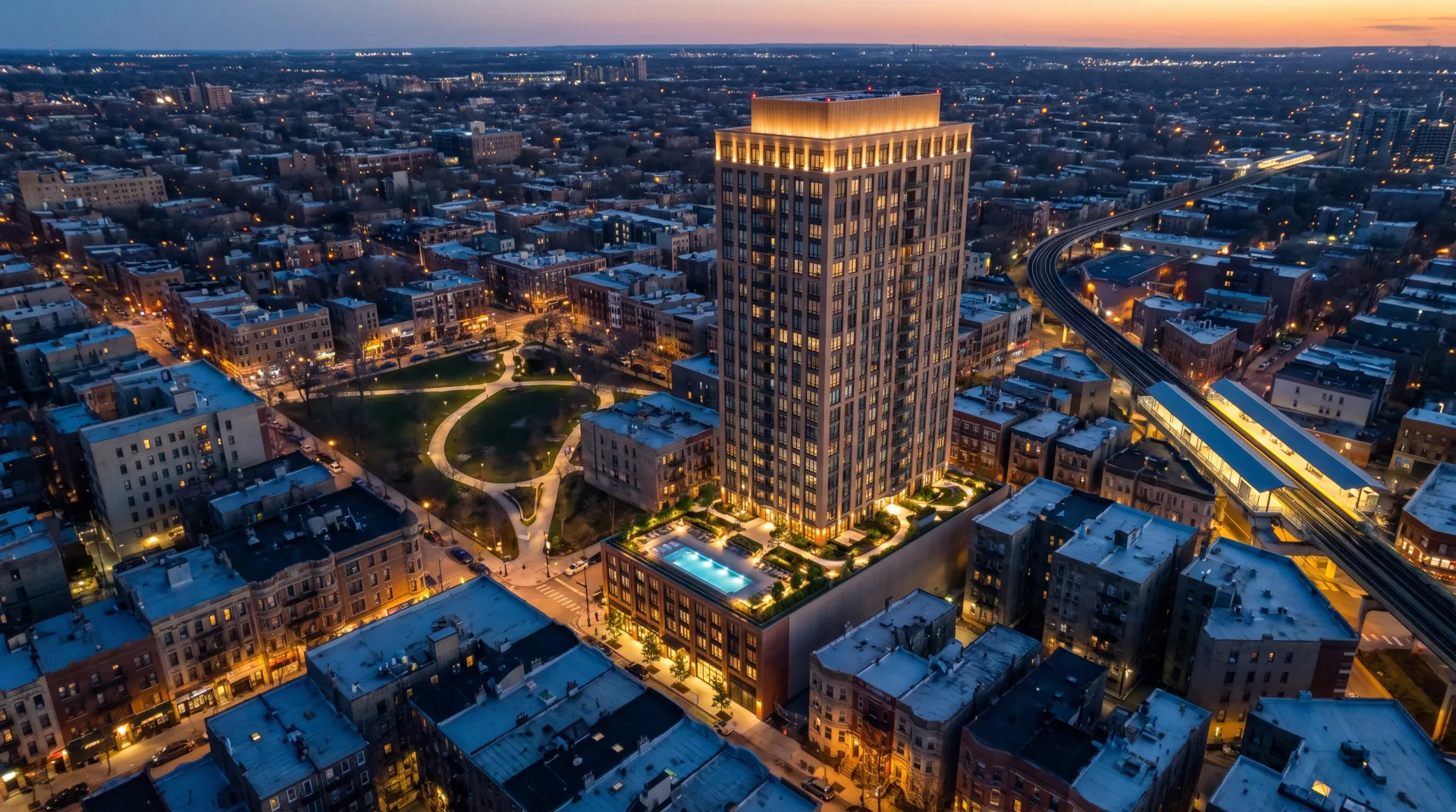 An aerial view at blue hour of a slender new residential tower with a lit crown and a pool deck, rising from a low-rise neighborhood near a park and an elevated rail station.