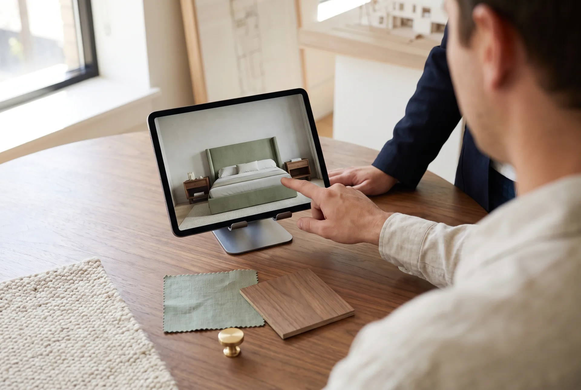 A tablet showing a rendered sage and walnut bedroom on a table among the matching fabric, veneer and brass samples, with a hand pointing at the screen.