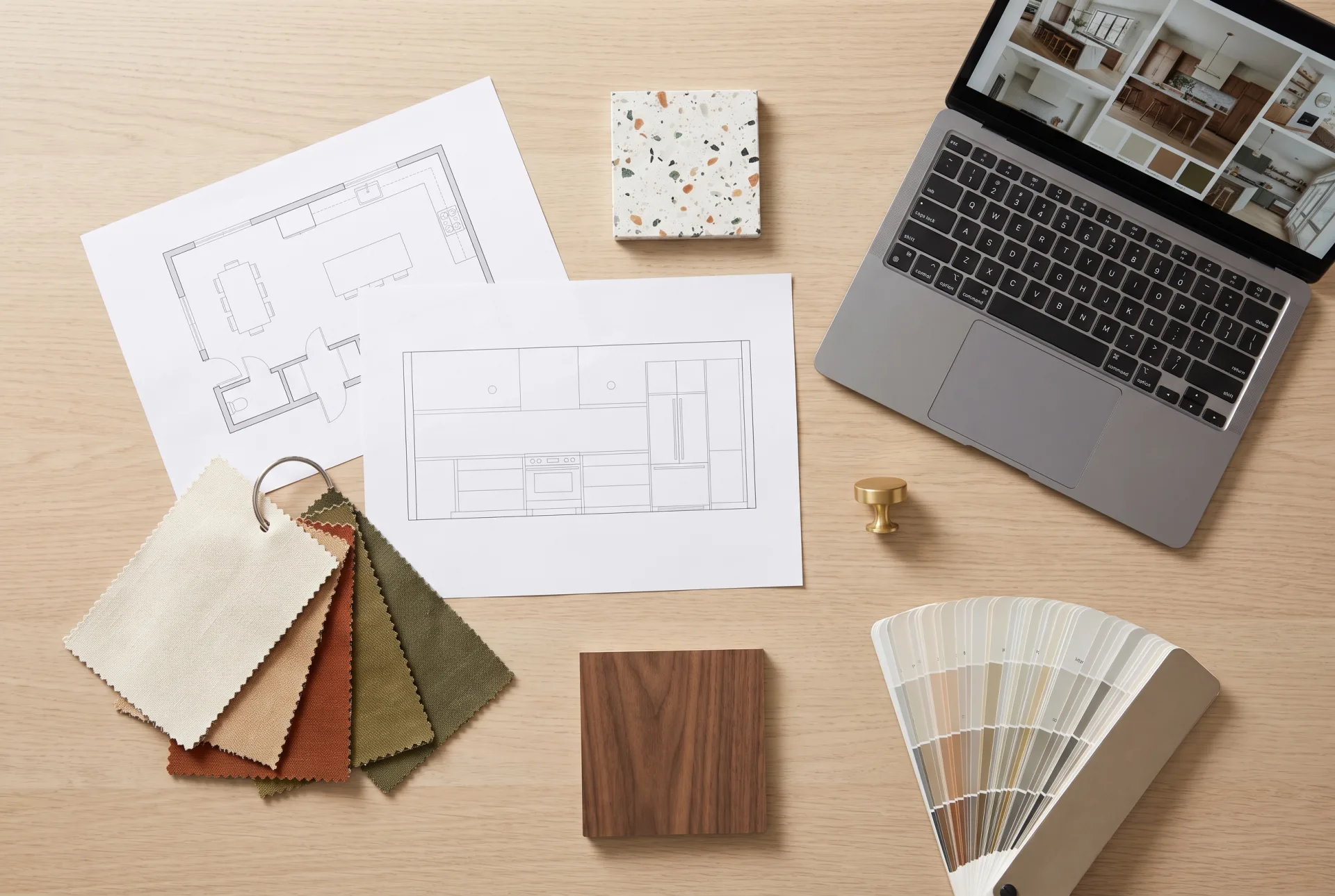 An overhead view of a desk with a printed floor plan, a kitchen elevation, fabric swatches, a terrazzo tile, a walnut sample, a brass knob and a laptop showing a mood board.