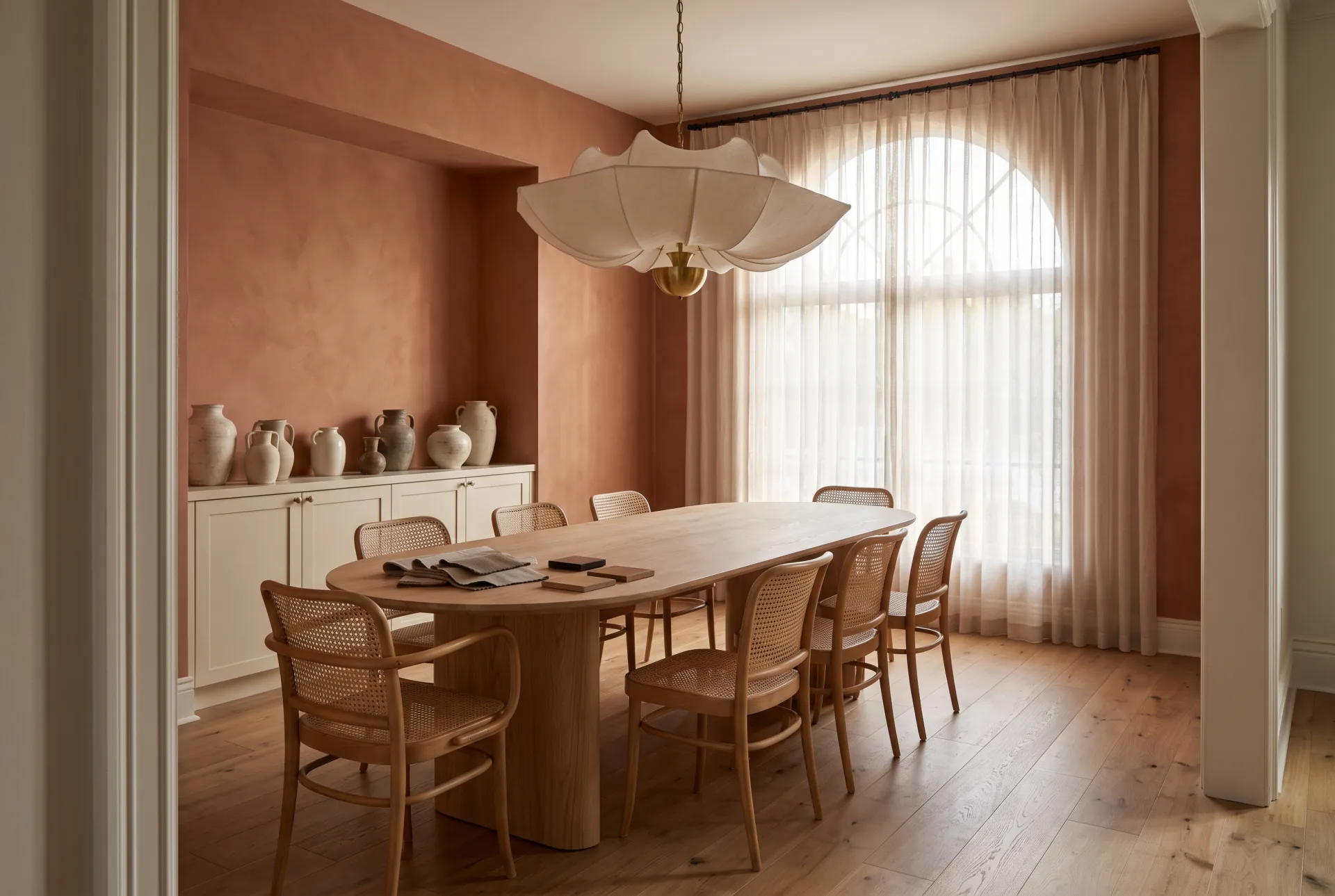 A terracotta dining room with an oval ash table, caned chairs and a paper chandelier, with fabric and wood samples laid on one end of the table.