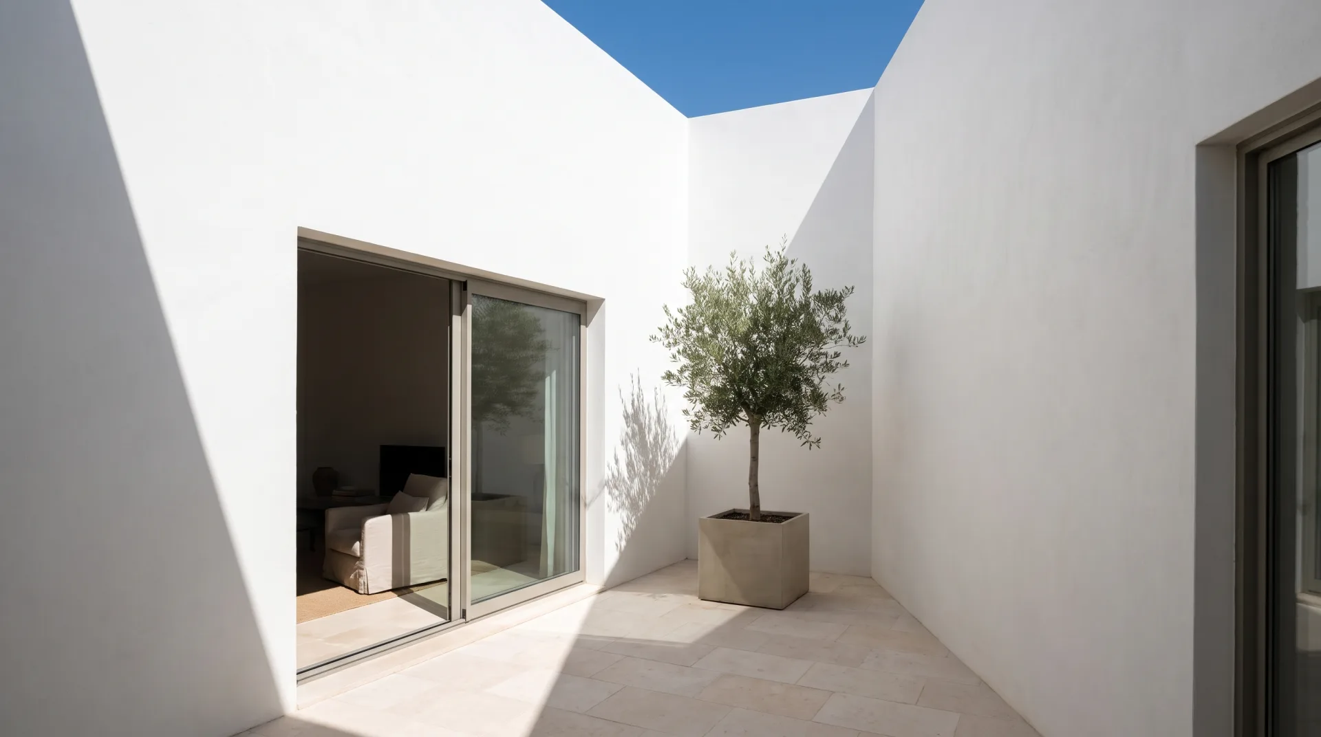 A white courtyard in midday sun where the shaded wall glows with light reflected from the sunlit wall and pale stone paving.