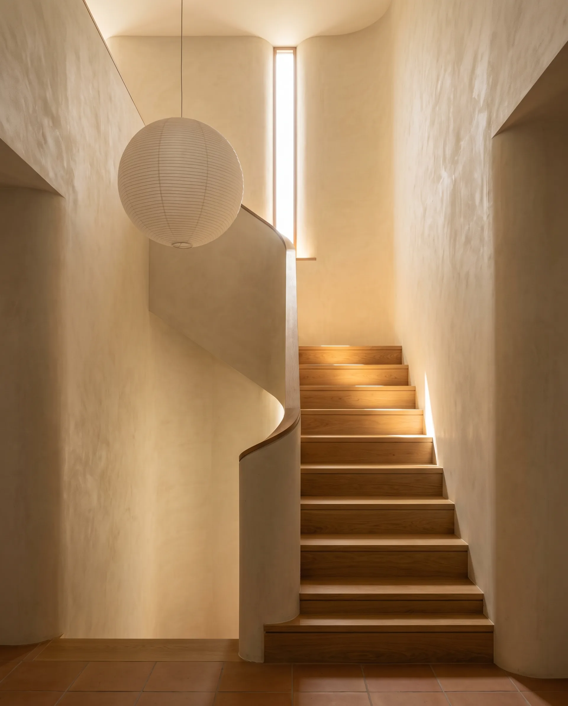 A double-height townhouse hall with a white-oak stair, lit by one shaft of sun and the warm light bouncing off plaster walls.