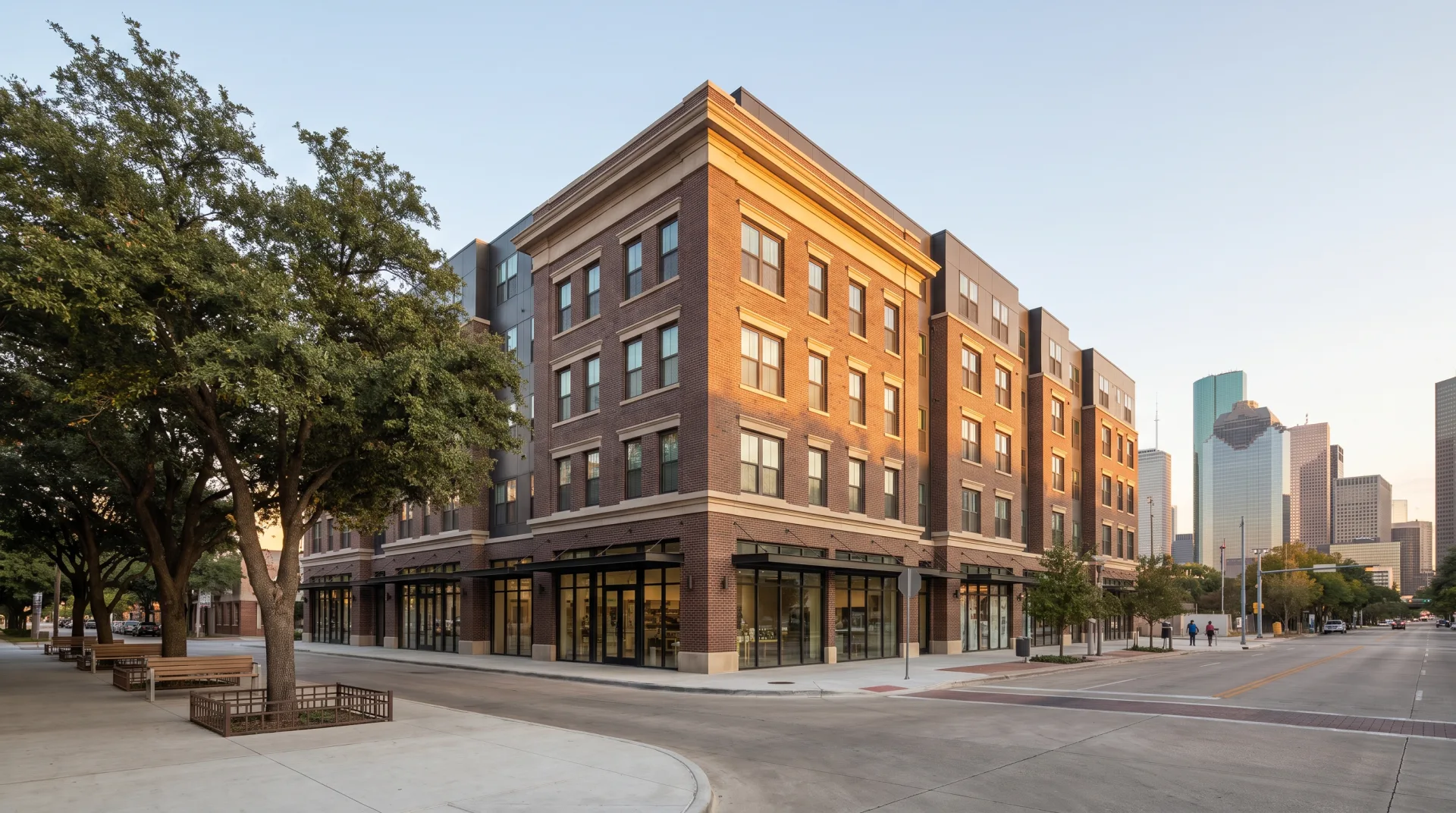 A six-story brick apartment building with cast-stone details and clear-glass shopfronts on a downtown-edge street corner in golden-hour light.