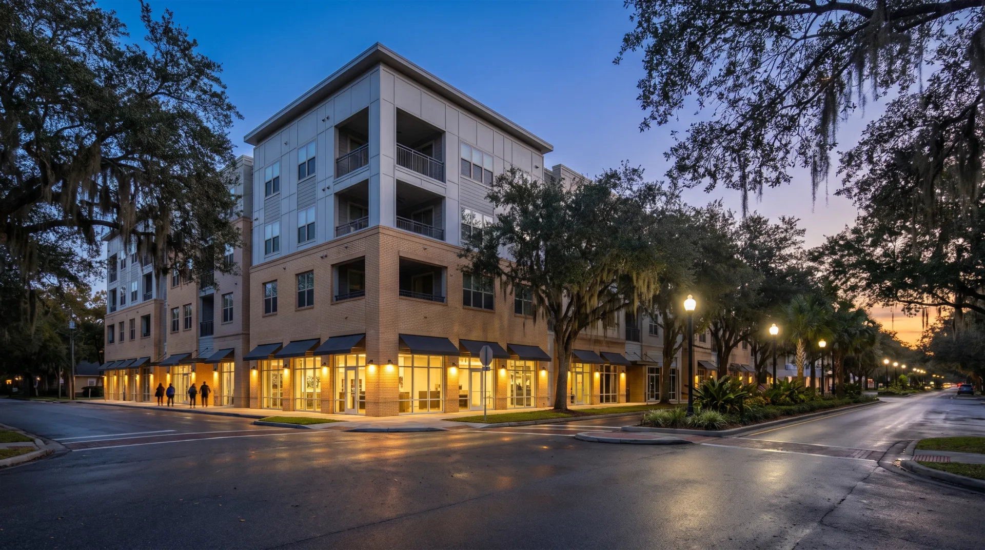 A new four-story brick and gray-panel mixed-use building on a boulevard of live oaks at dusk, its ground-floor shops lit.