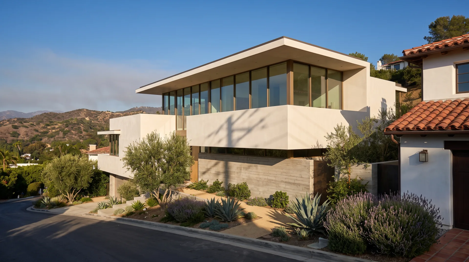 A new white stucco and glass house stepping down a Los Angeles hillside with olive trees and drought-tolerant planting in late-afternoon sun.
