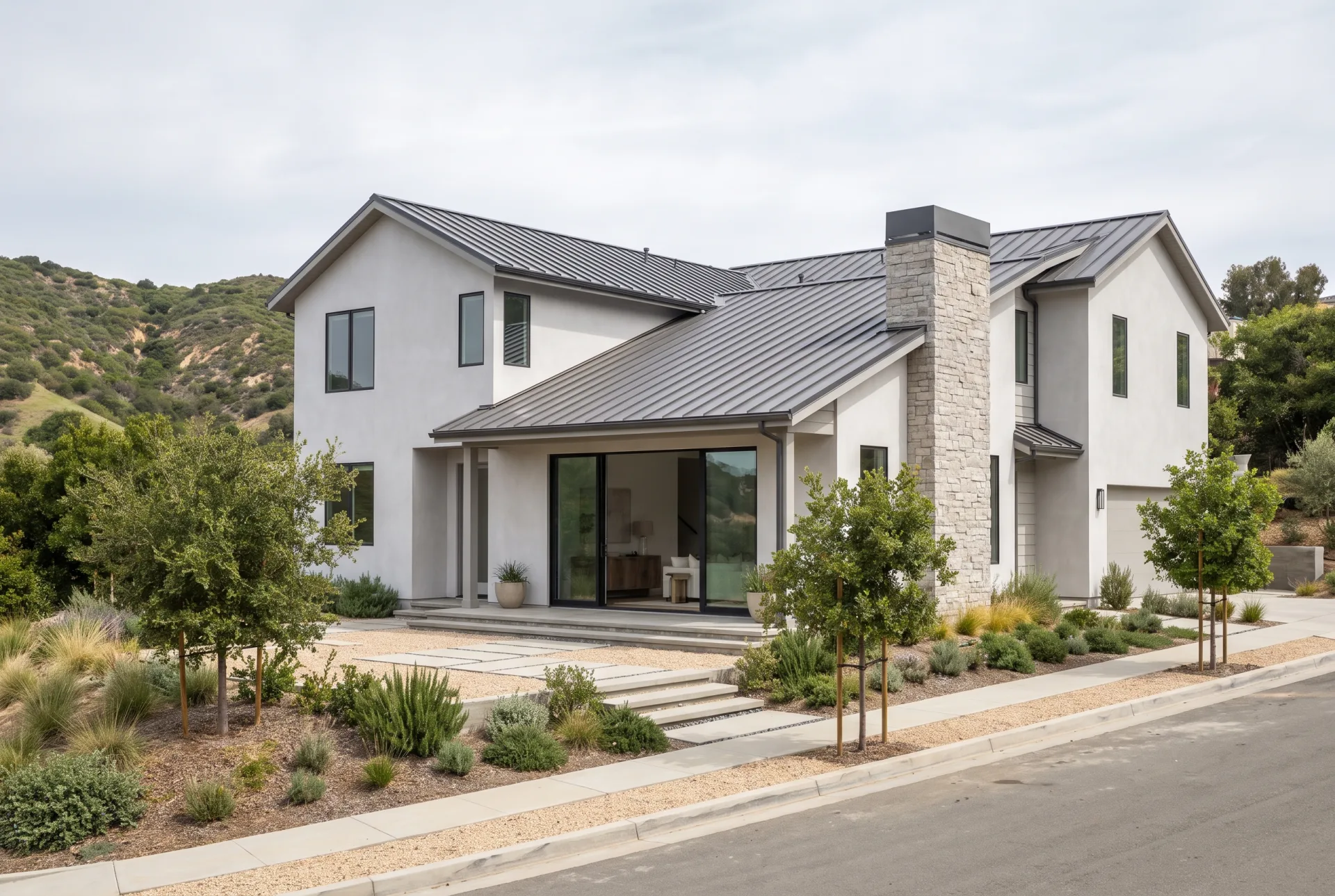 A newly built two-story plaster house with a metal roof and native planting on a sloping lot in a green coastal canyon neighborhood.