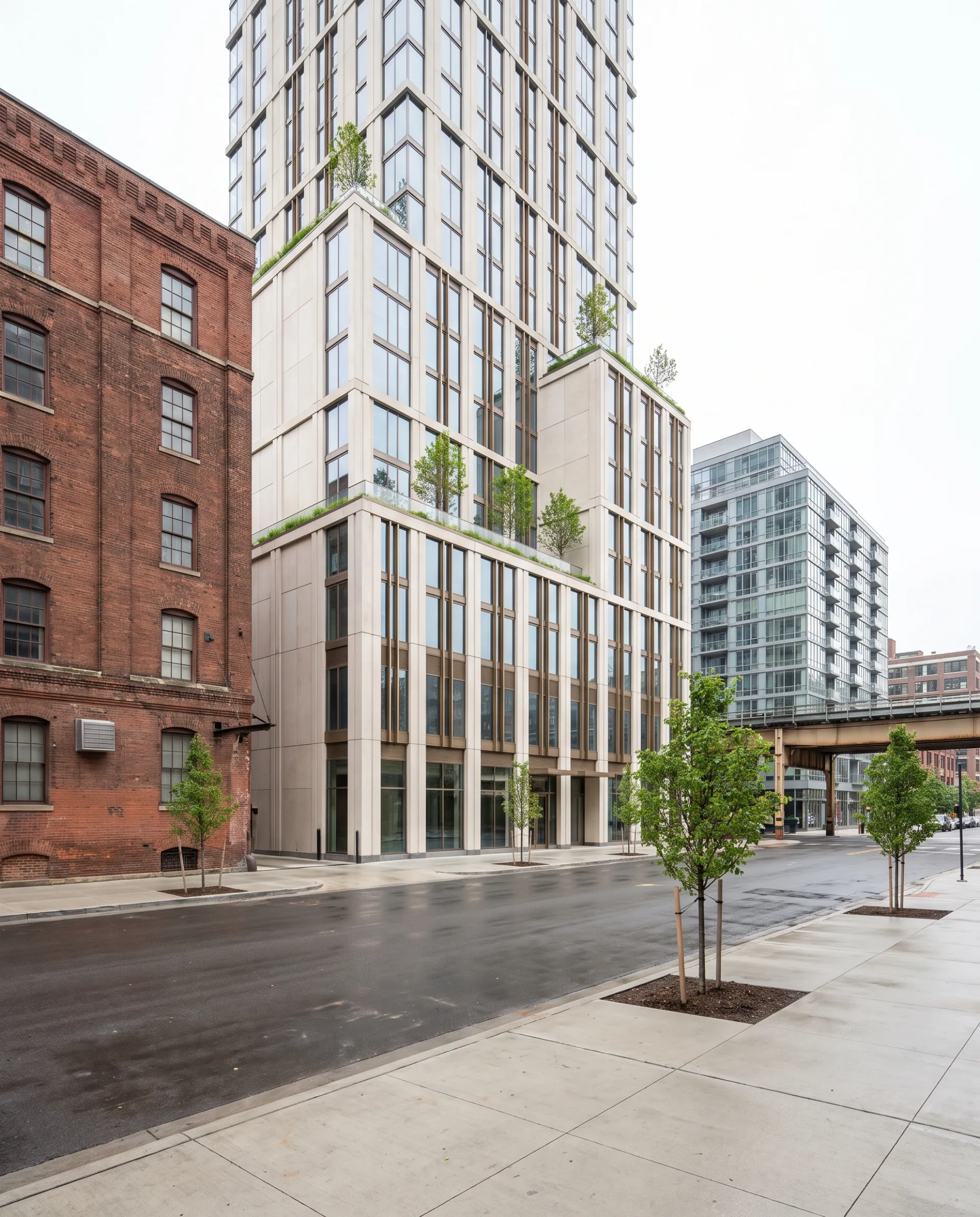A new 24-story condo tower with bronze fins shown at true scale between a brick loft building and a glass rental, with young street trees and an elevated rail line.