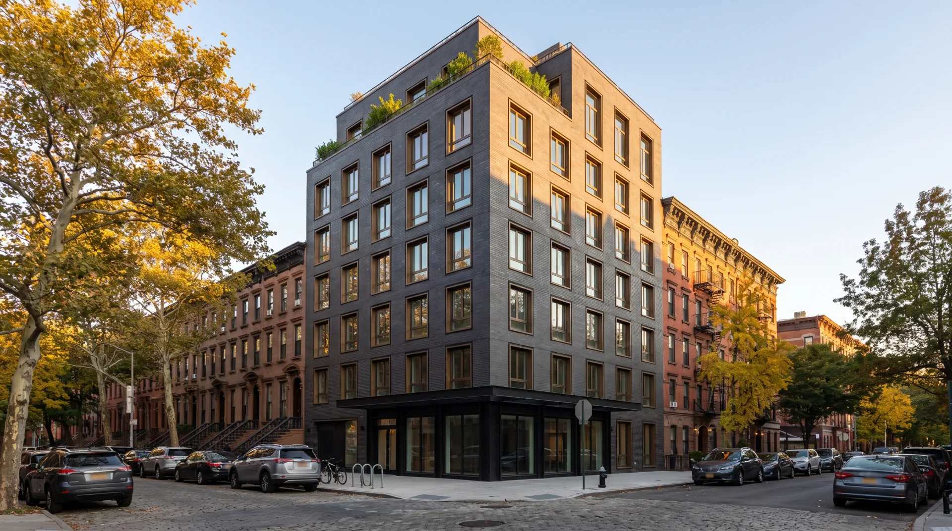 A new eight-story charcoal brick apartment building on a Brooklyn corner between brownstone rowhouses and a red-brick tenement, in late-afternoon autumn sun.