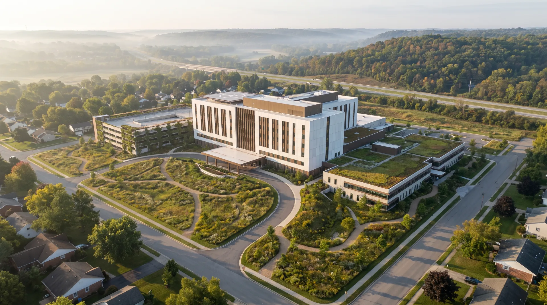 A high aerial view of a new hillside medical campus with a white main building, a green-roofed wing, a parking structure and meadow paths, among suburban streets in morning haze.