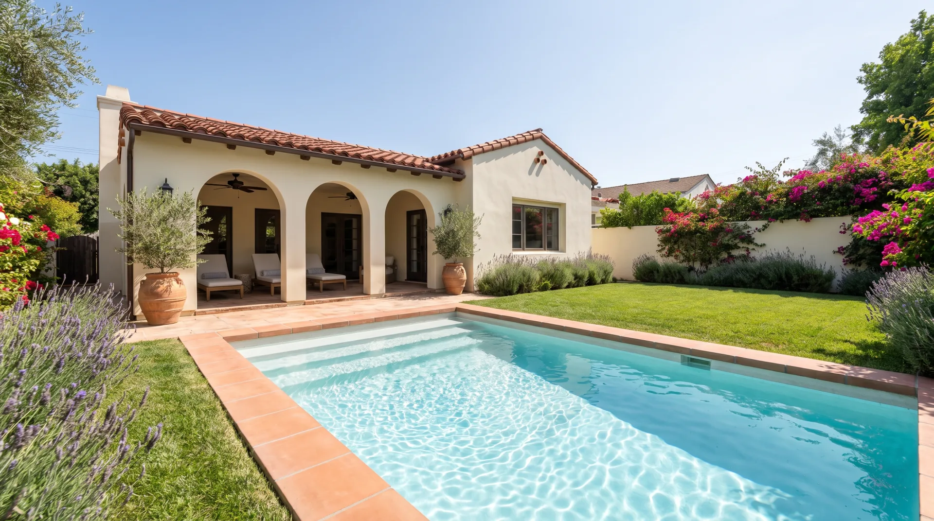 A backyard pool with terracotta coping in front of a Spanish-revival house with an arched loggia and potted olive trees.
