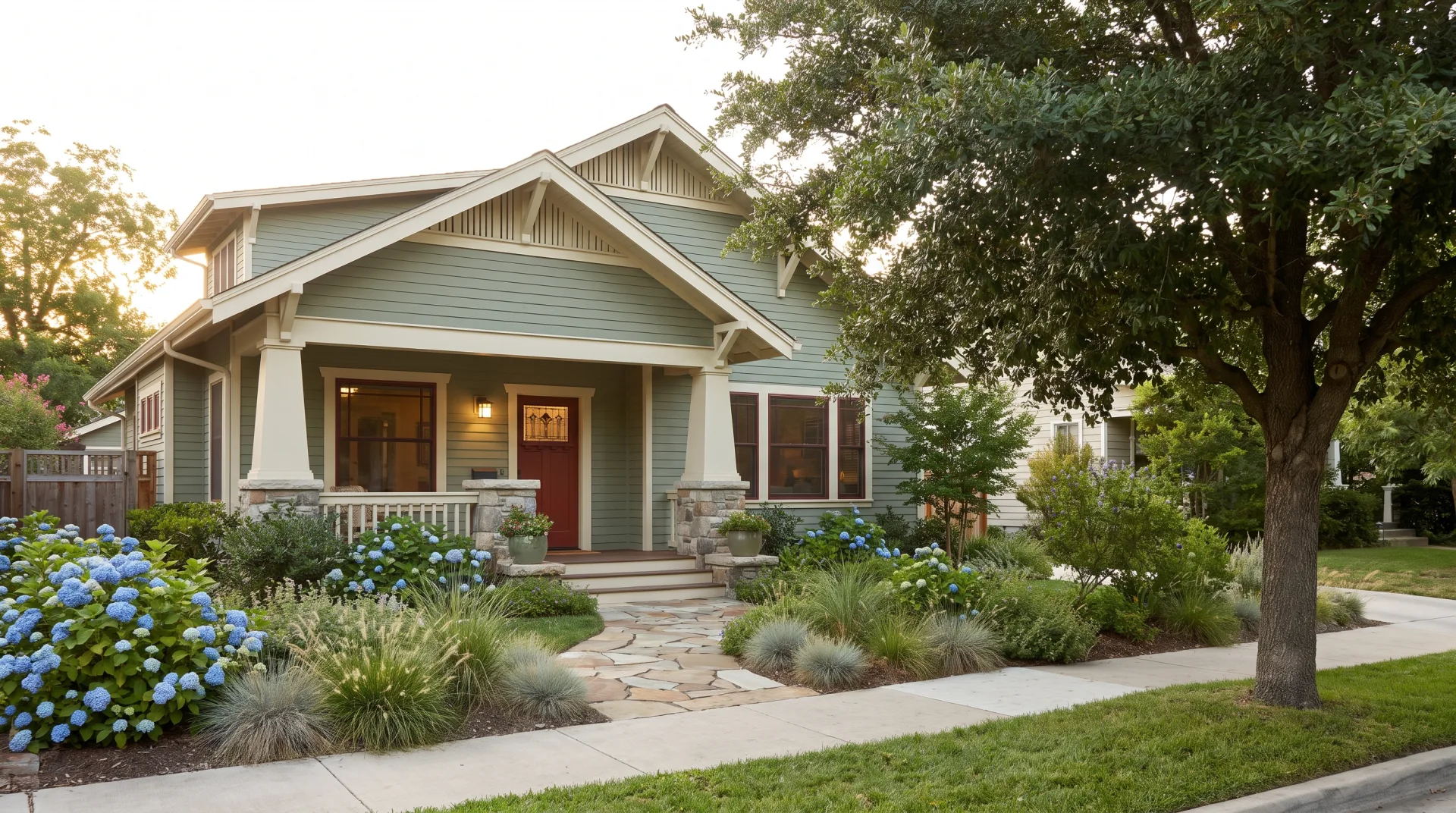 A new Craftsman house with sage green siding, a deep stone-columned porch and a red front door behind a planted front yard.