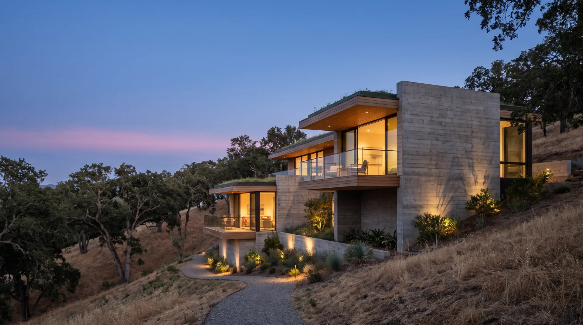 A concrete hillside house stepping down a slope at dusk, its glass walls glowing warm against a blue sky.