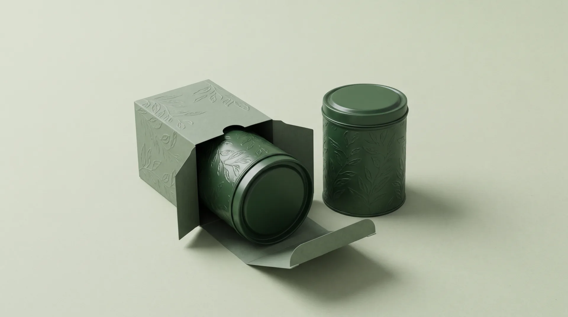 A forest green tea tin with an embossed botanical pattern beside its open paper carton on a pale sage backdrop.