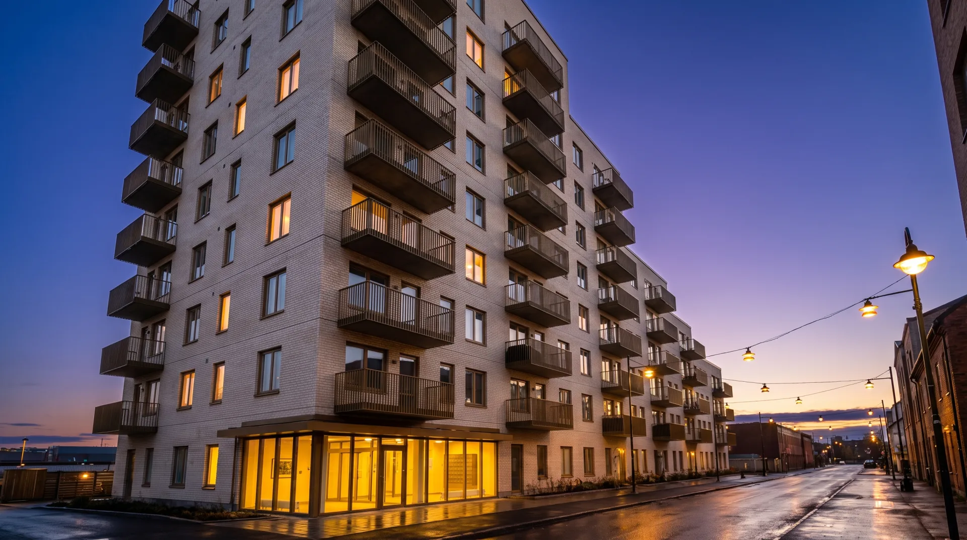 The same pale brick apartment building at dusk with warm lit windows and a glowing lobby under a deep blue sky.
