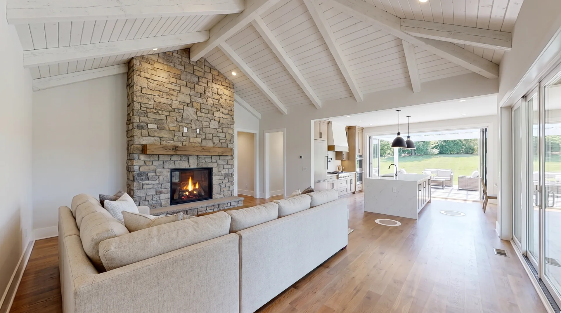 A rendered model home great room with a vaulted beamed ceiling, stone fireplace and open kitchen, with two round navigation hotspots on the floor.
